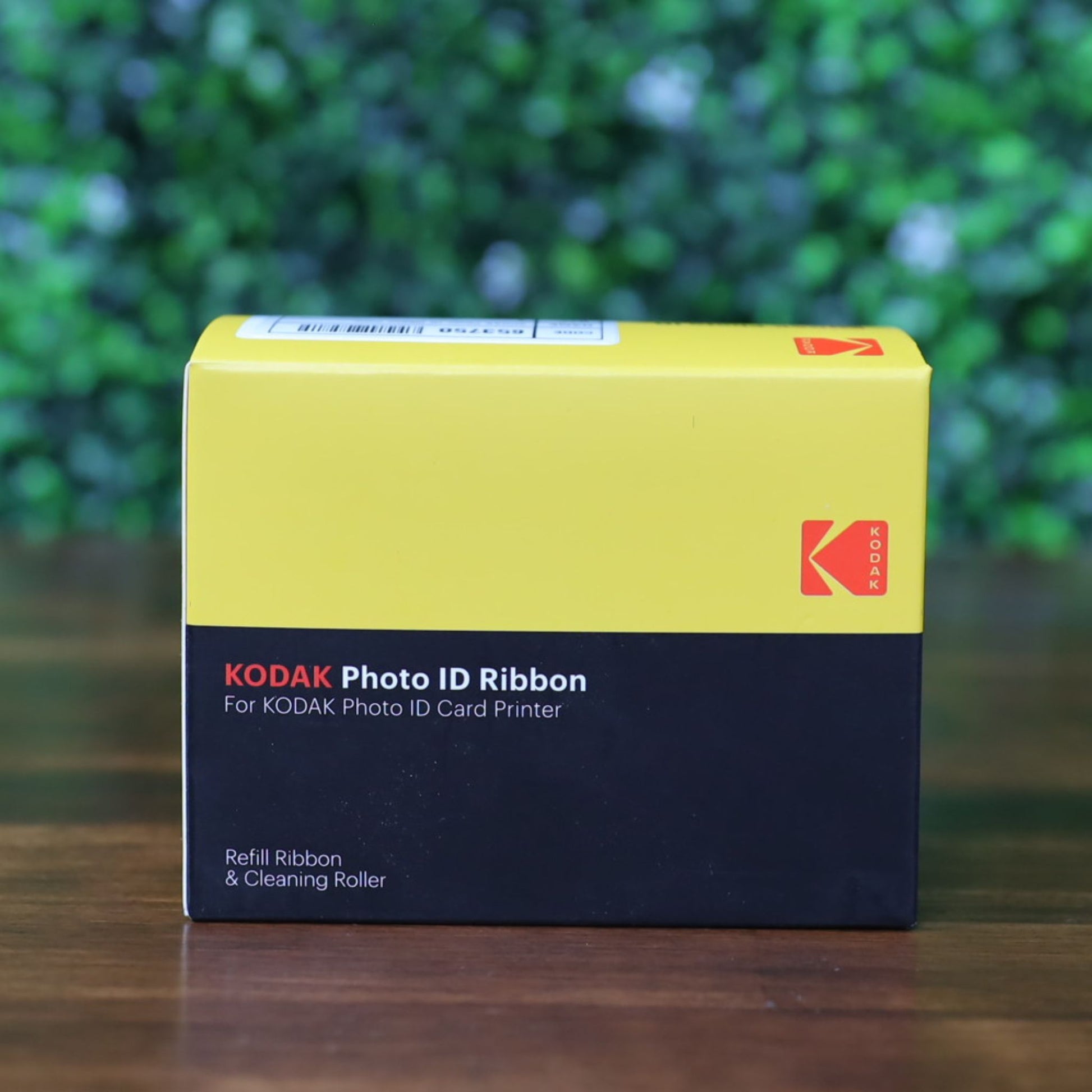 A KODAK 653750 Photo ID Color Ribbon for KODAK ID100S and ID200S card printers (YMCKO, 100 prints) in its yellow and black retail box with barcode label, displayed on a wooden surface with a blurred green leafy background.  Photographed and sold by Specialist ID, an authorized Magicard dealer.