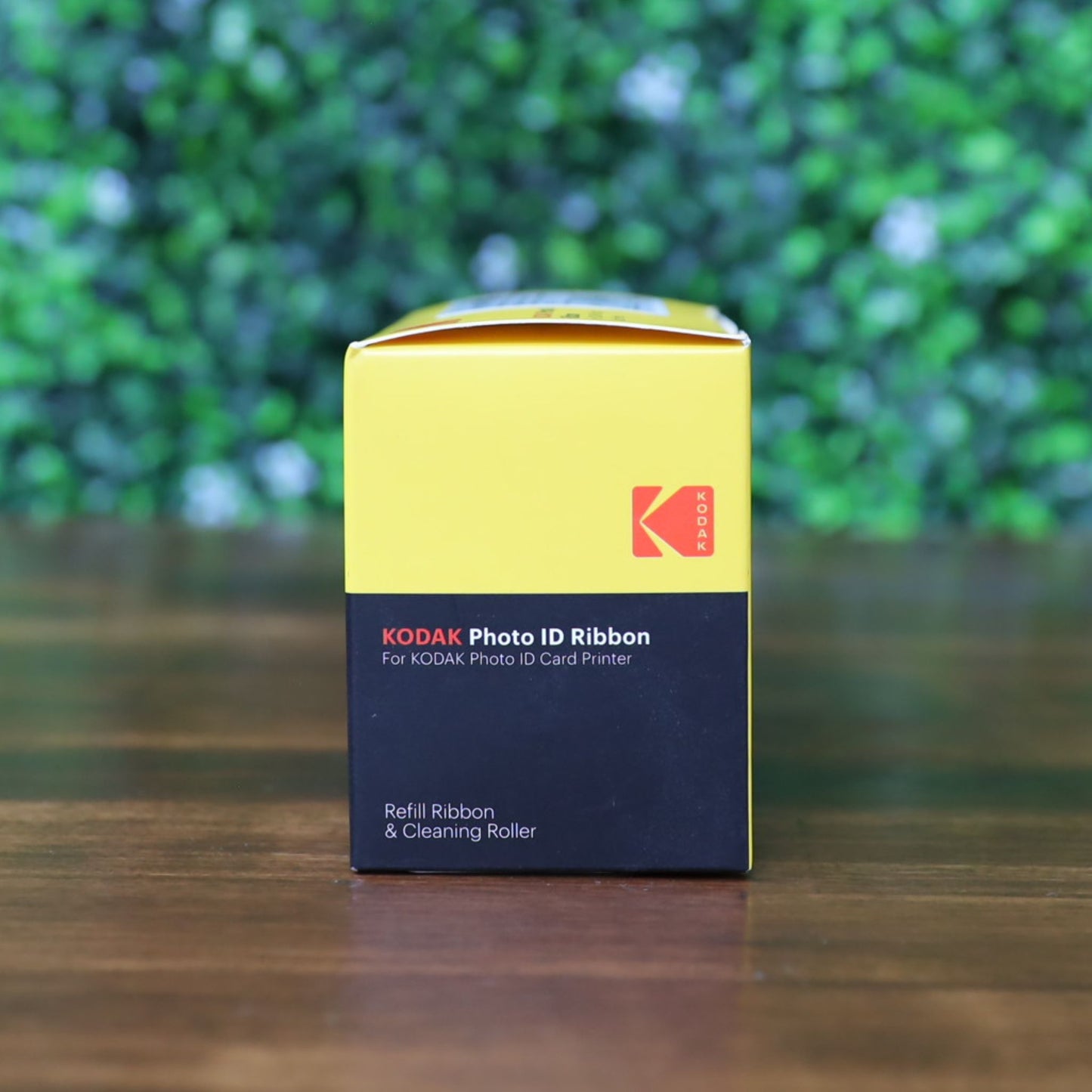A box of KODAK 653750 Photo ID Color Ribbon for KODAK ID100S and ID200S card printers (YMCKO, 100 prints) in yellow and black retail packaging, displayed on a wooden surface with a blurred green leafy background. Photographed and sold by Specialist ID, an authorized Magicard dealer.