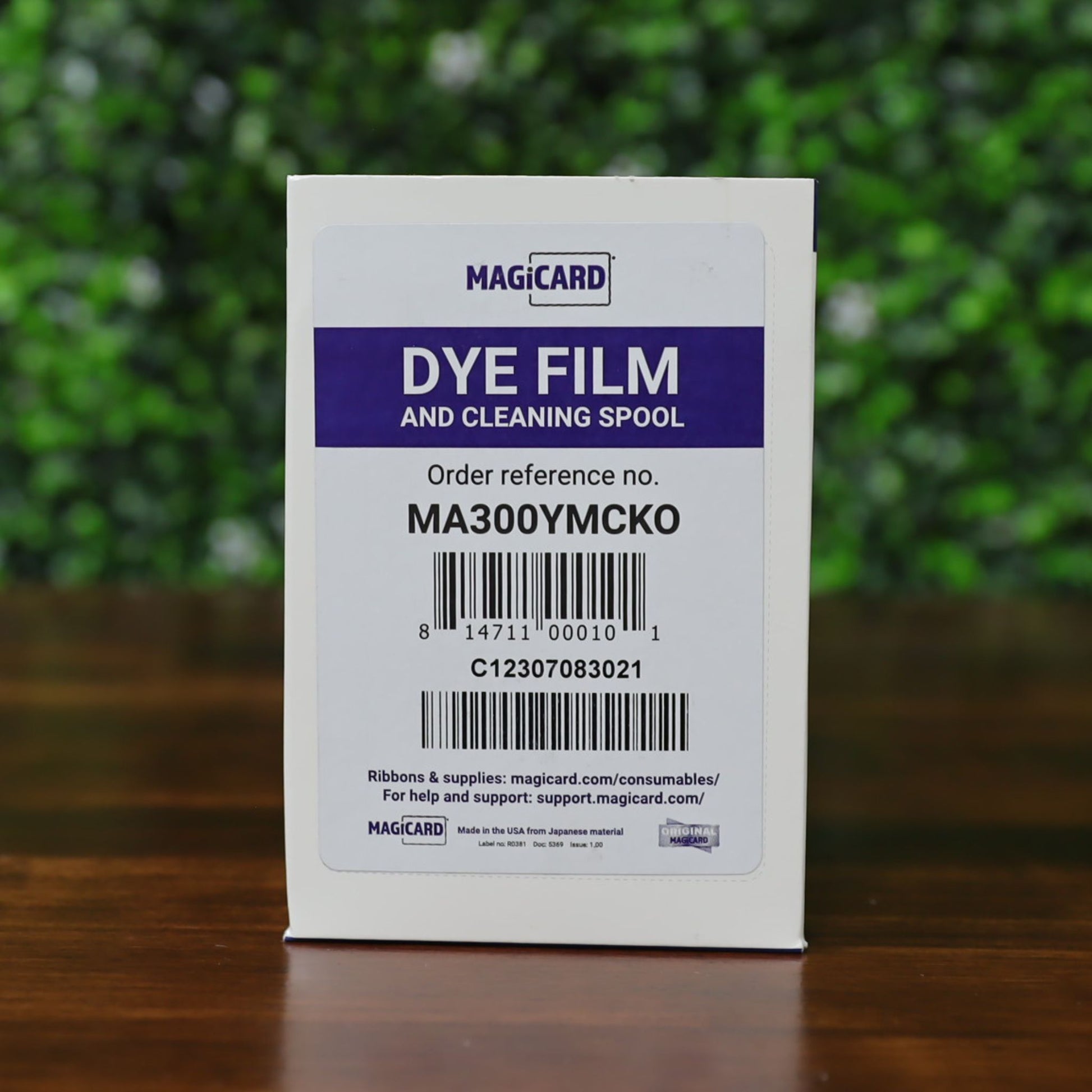 A box labeled "Magicard MA300YMCKO YMCKO Color Ribbon - 300 Prints" with order reference MA300YMCKO sits on a wooden surface, set against a green blurred background. Photographed and sold by Specialist ID, an authorized Magicard dealer.