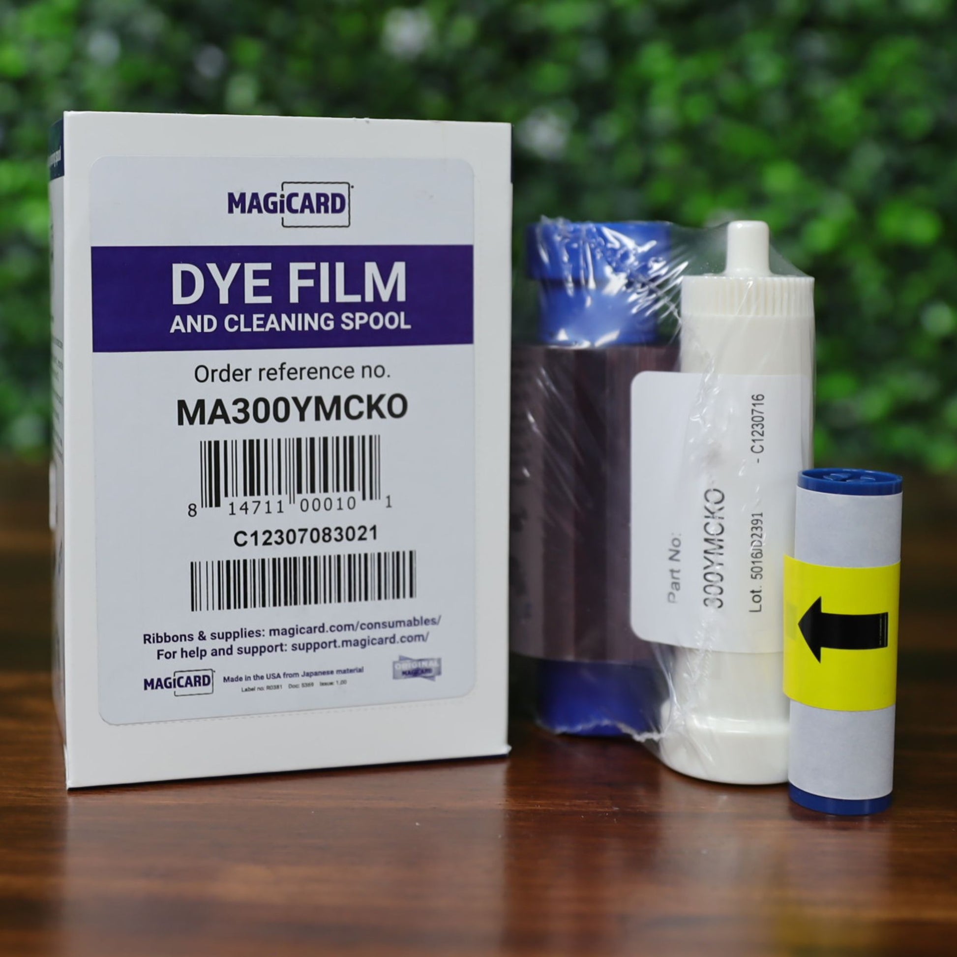 Magicard MA300YMCKO Color Ribbon – 300 prints, boxed with color ribbon cartridge and cleaning spools, displayed on a wooden surface with a blurred green leafy background. Photographed and sold by Specialist ID, an authorized Magicard dealer.