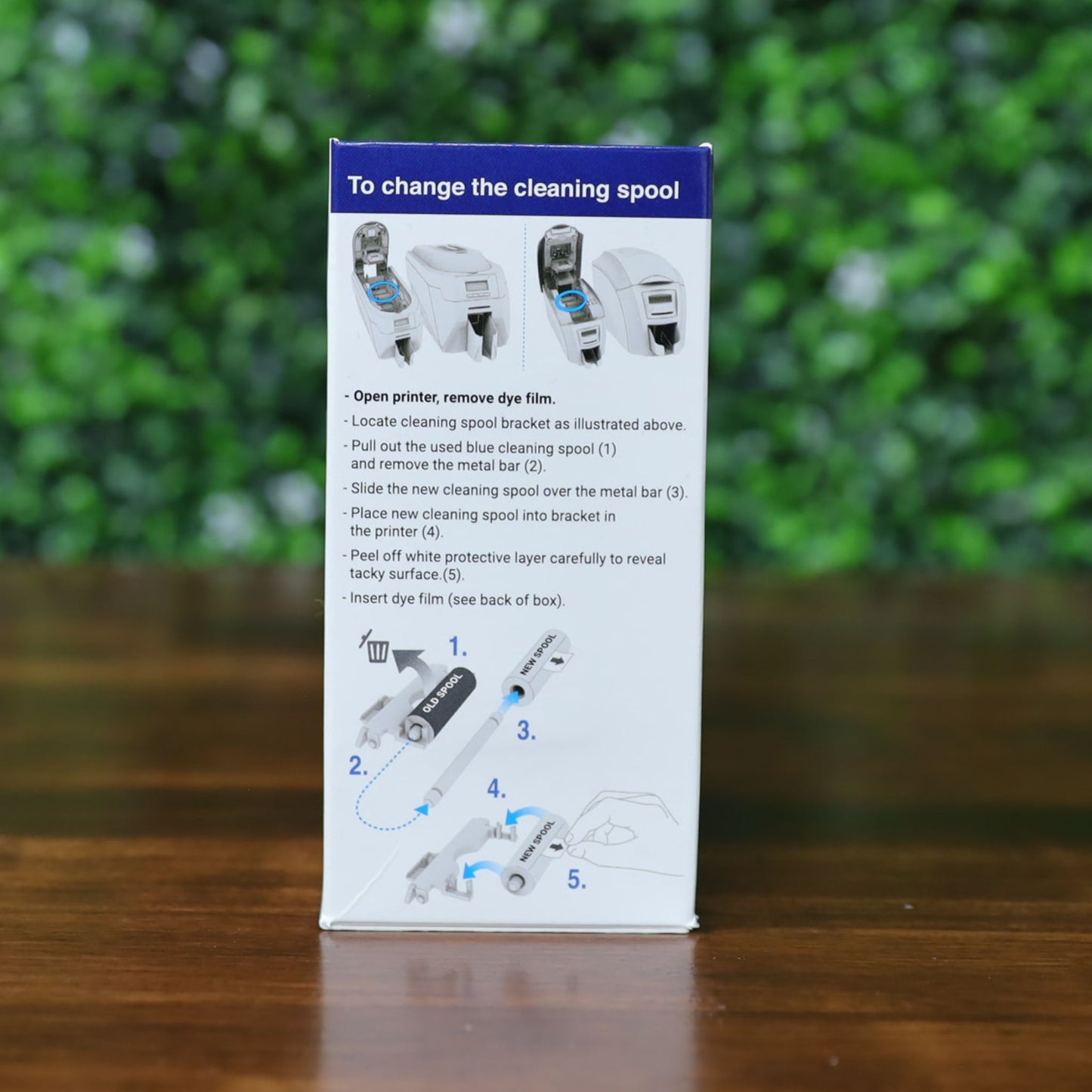 Box displaying step-by-step instructions and illustrations for changing the cleaning spool of the Magicard MA300YMCKO YMCKO Color Ribbon - 300 Prints, set against a blurred green background. No SEO keywords can be extracted from CSS code alone. Photographed and sold by Specialist ID, an authorized Magicard dealer.