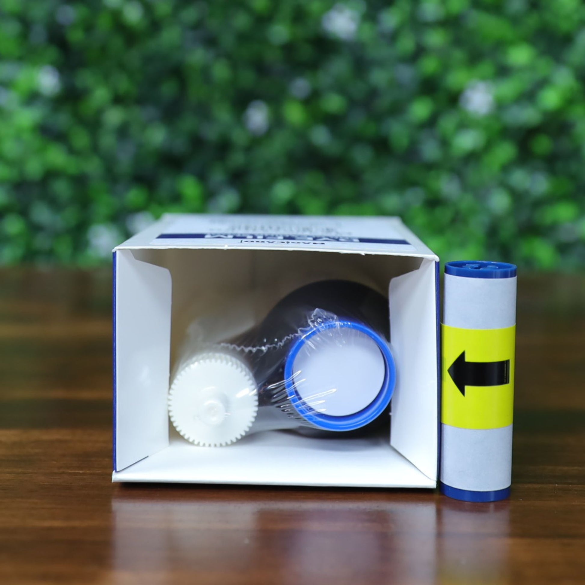 An open box of Magicard MA300YMCKO YMCKO Color Ribbon (300 prints), showing two bottles—one with a blue cap, one with a white cap—rests on wood next to a rolled sheet featuring a yellow arrow label. Photographed and sold by Specialist ID, an authorized Magicard dealer.