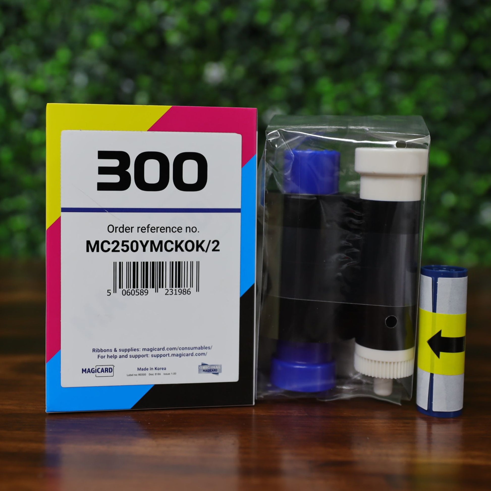 A Magicard MC250YMCKOK Ribbon box for 250 dual-sided full-color prints, containing two rolls, rests on a wooden surface with a leafy background—ideal for vibrant, reliable card printing.