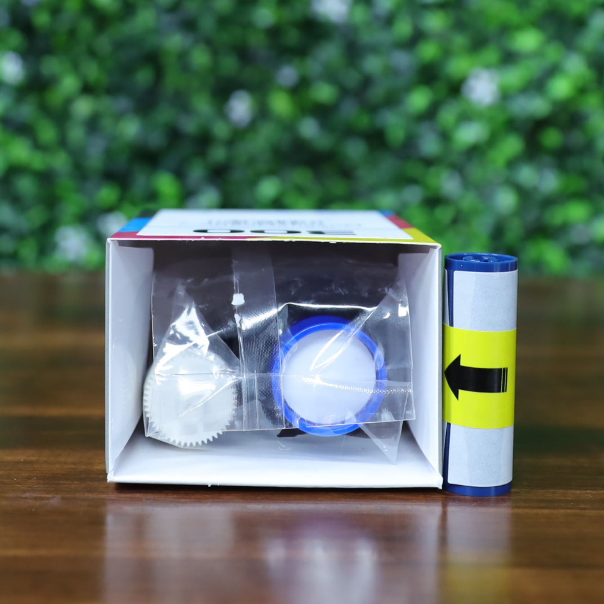 A Magicard MC250YMCKOK Ribbon (for 250 dual-sided full-color prints) with a blue film roll and cleaning roller visible inside an open box lying on its side, displayed on a wooden surface with leafy greenery in the background. Photographed and sold by Specialist ID, an authorized Magicard dealer.