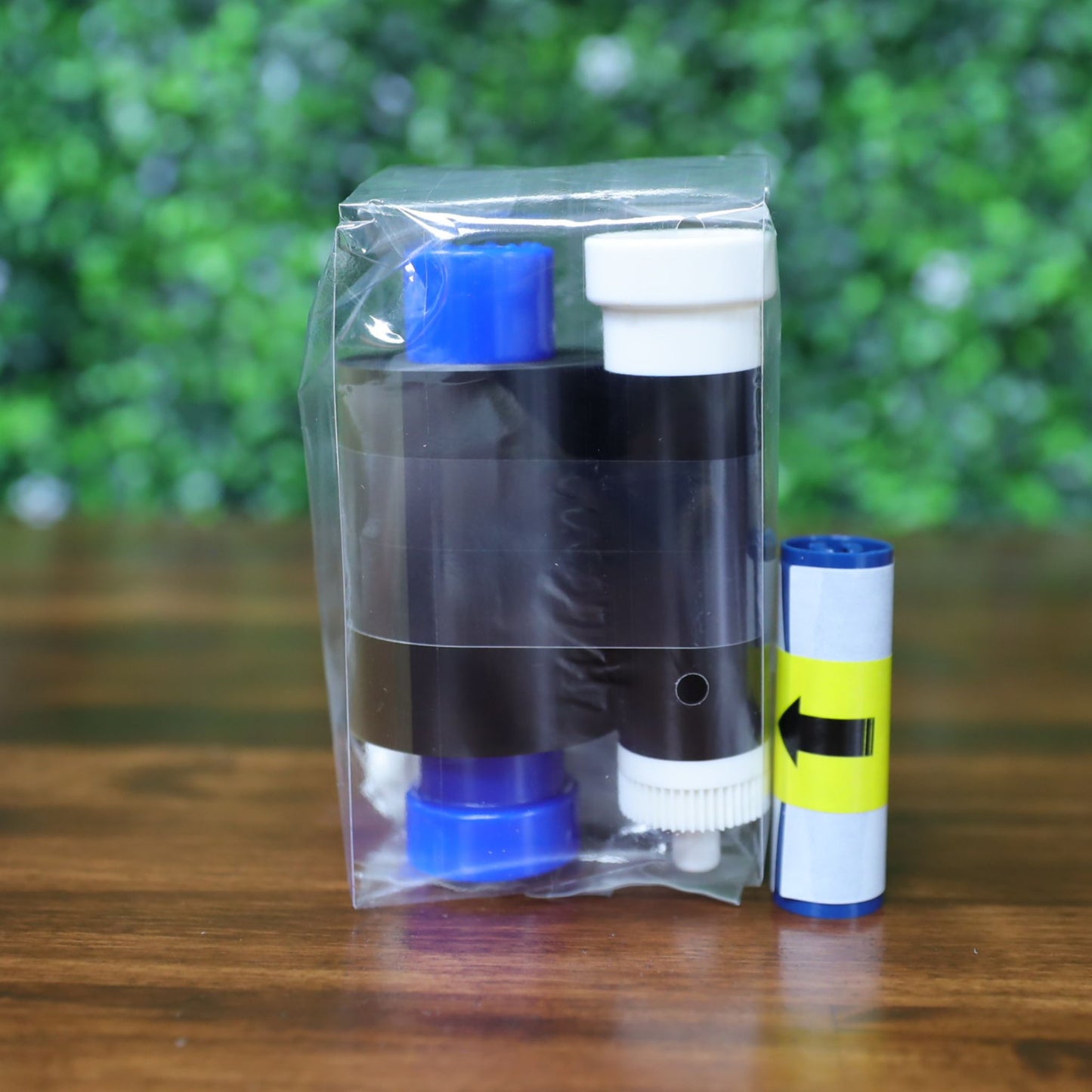 A clear plastic pouch holds a Magicard MC250YMCKOK Ribbon for 250 dual-sided full-color prints, with a black and blue refill cartridge and a small rolled blue and yellow item, displayed on wood against a green background.