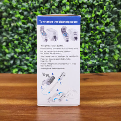 The Magicard MA1000K-Black Monochrome K Ribbon - 1000 Prints packaging features illustrated, step-by-step ribbon change instructions on a leafy green background and wood surface—ideal for your monochrome ID card printing needs.