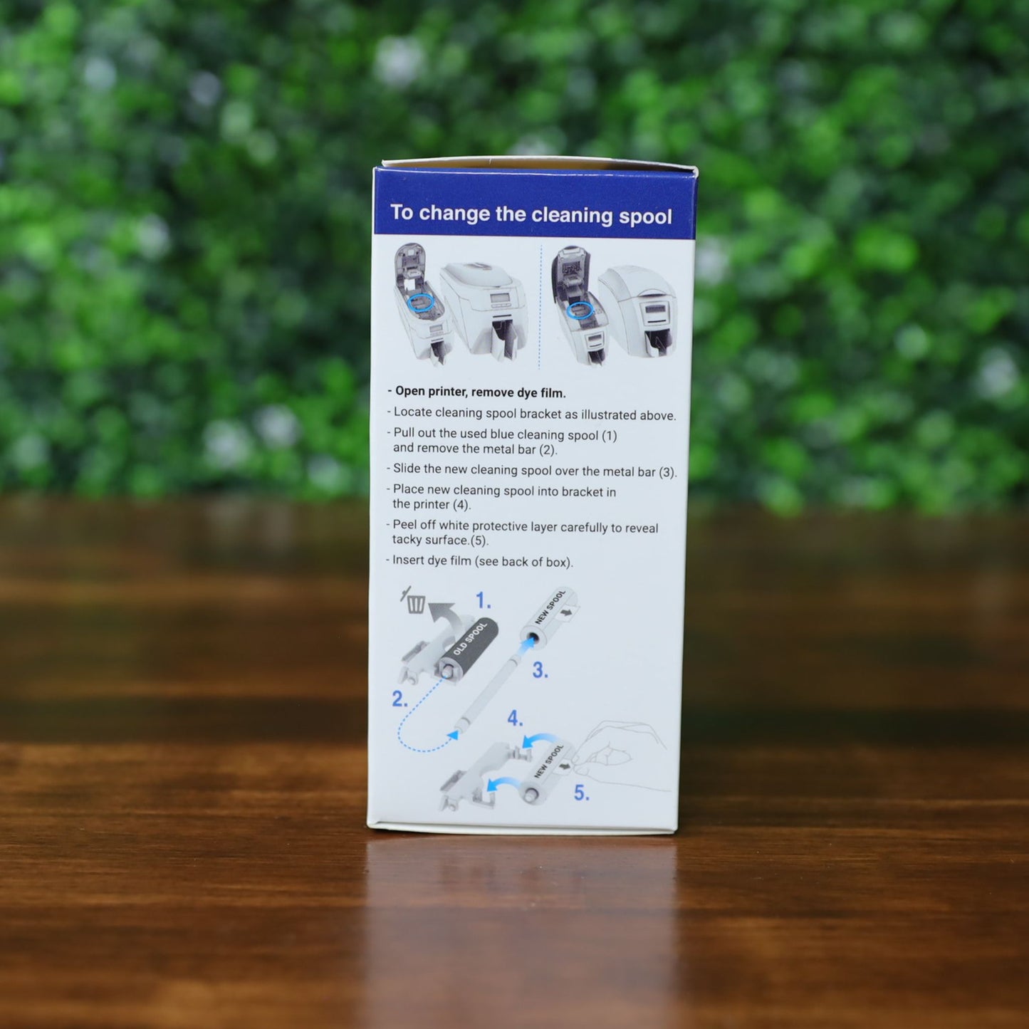 Box displaying step-by-step printed instructions and diagrams for changing the cleaning spool of the Magicard MA1000K GOLD Gold Ribbon – 1000 Prints, set on a wooden surface with a green leafy background. No product description; only CSS code shown. Photographed and sold by Specialist ID, an authorized Magicard dealer.
