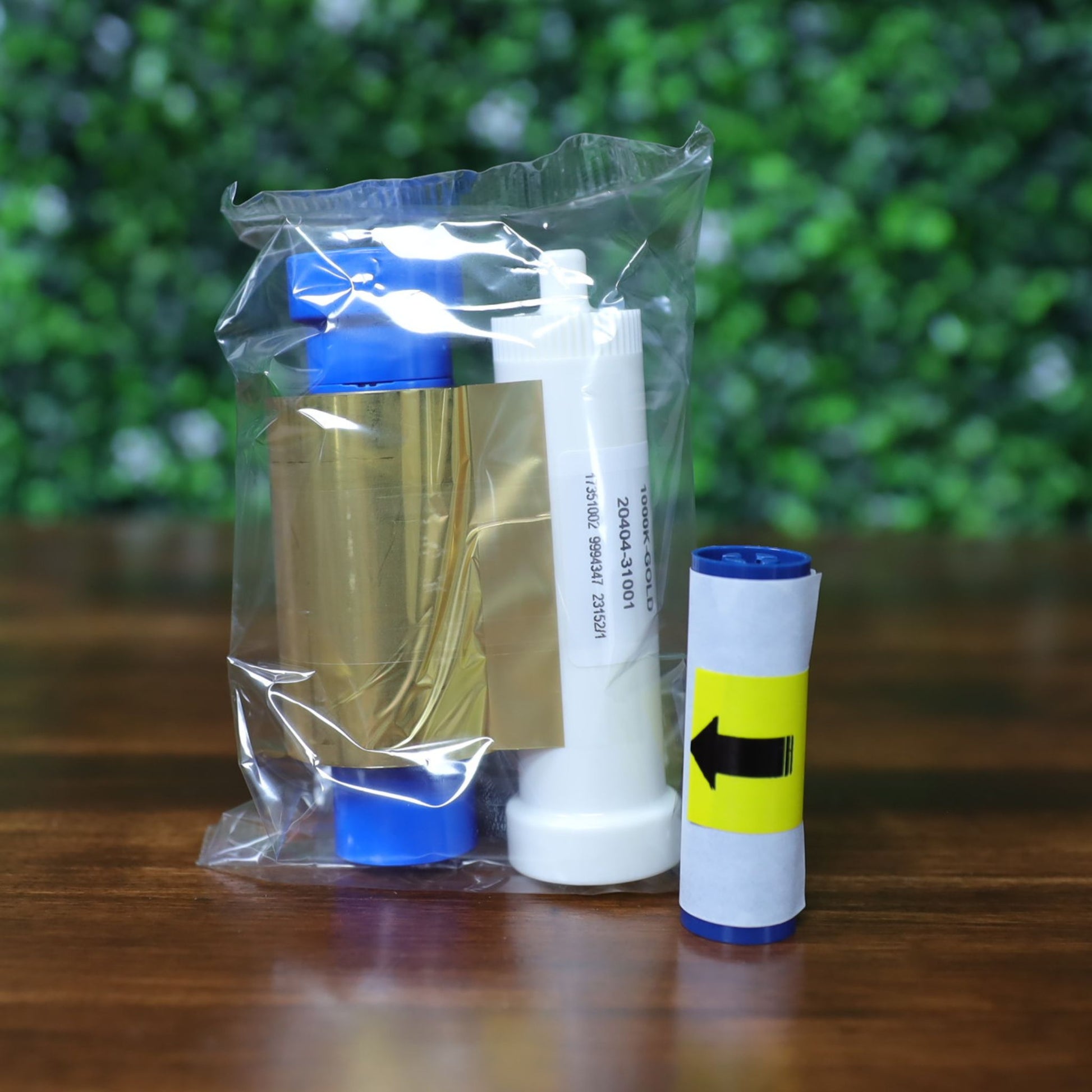 A sealed plastic pack contains a Magicard MA1000K GOLD Ribbon (1000 prints) in blue and white, plus a separate rolled paper with a yellow arrow label, displayed on wood with a green blurred background. Photographed and sold by Specialist ID, an authorized Magicard dealer.