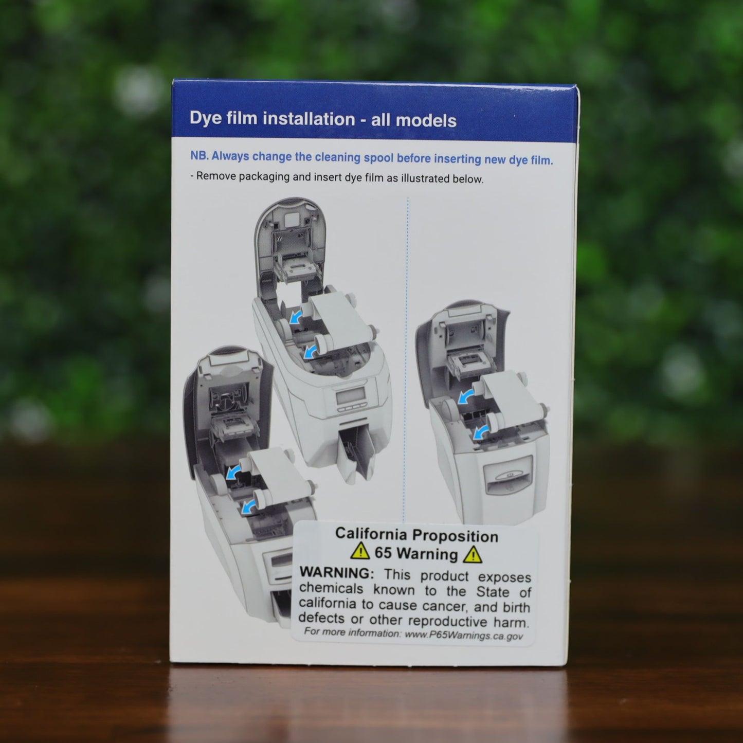 Back of Magicard MA1000K-Scratch scratch-off ribbon box showing step-by-step illustrated instructions for replacing the cleaning spool, with text and diagrams, displayed on a wood surface with green leafy background, photographed and sold by Specialist ID, an authorized Magicard dealer.