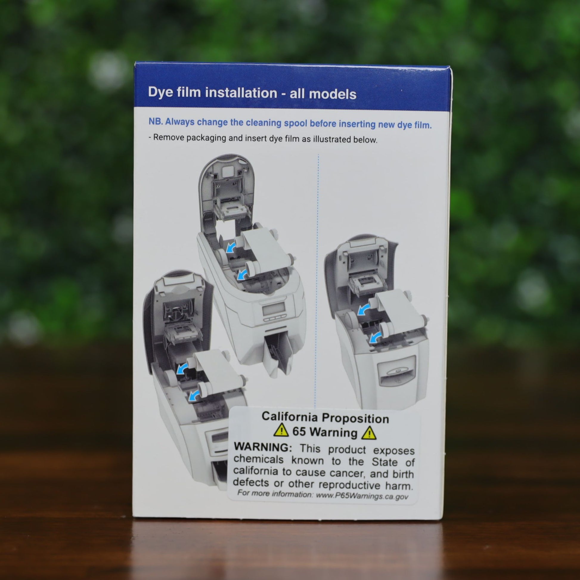 Back of Magicard MA1000K-Scratch scratch-off ribbon box showing step-by-step illustrated instructions for replacing the cleaning spool, with text and diagrams, displayed on a wood surface with green leafy background, photographed and sold by Specialist ID, an authorized Magicard dealer.