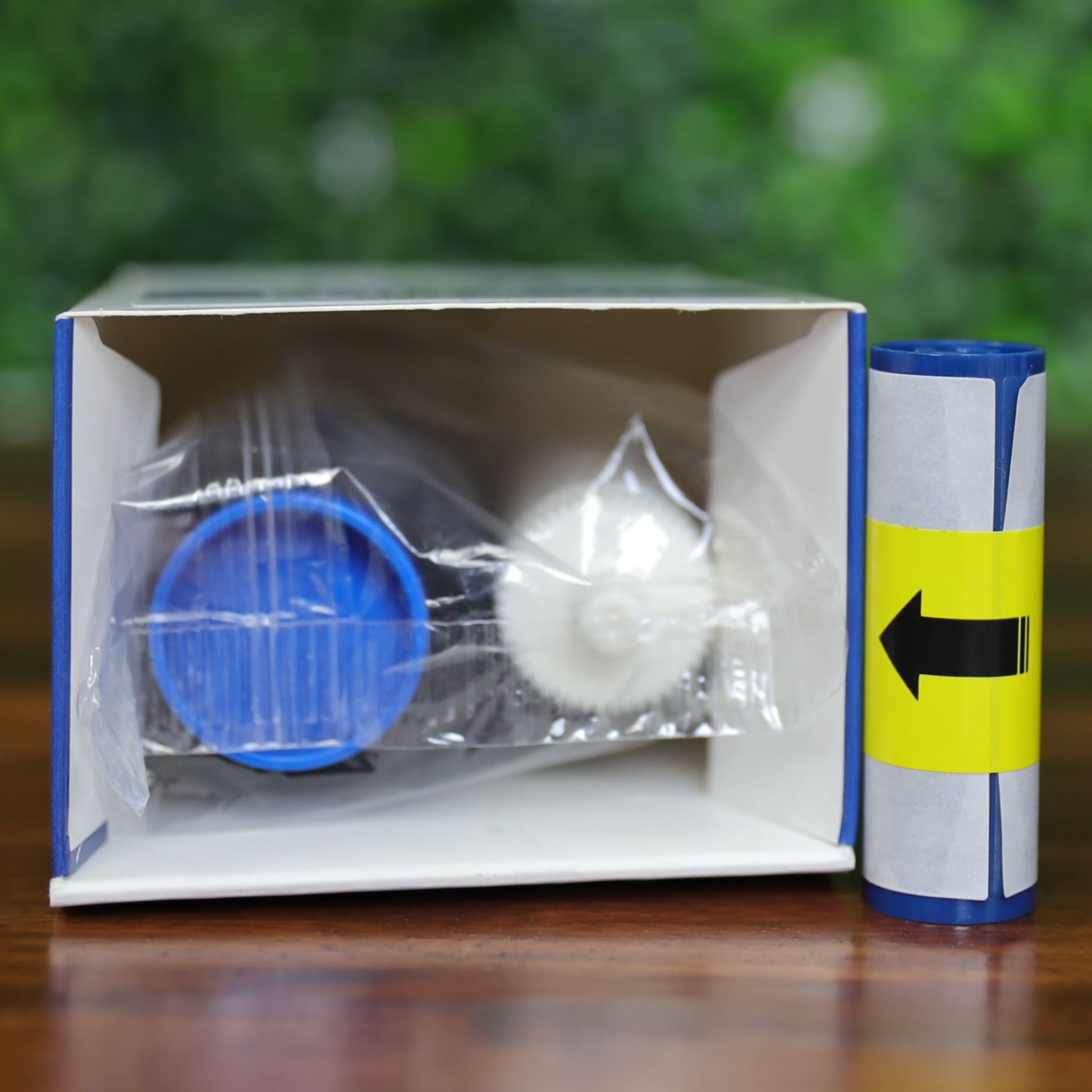 Top view of an open Magicard MA1000K-Scratch scratch-off ribbon kit showing a blue-capped ribbon with wavy gray pattern, white cleaning spool, and yellow label with black arrow, on a wood surface with green leafy background, photographed and sold by Specialist ID, an authorized Magicard dealer.