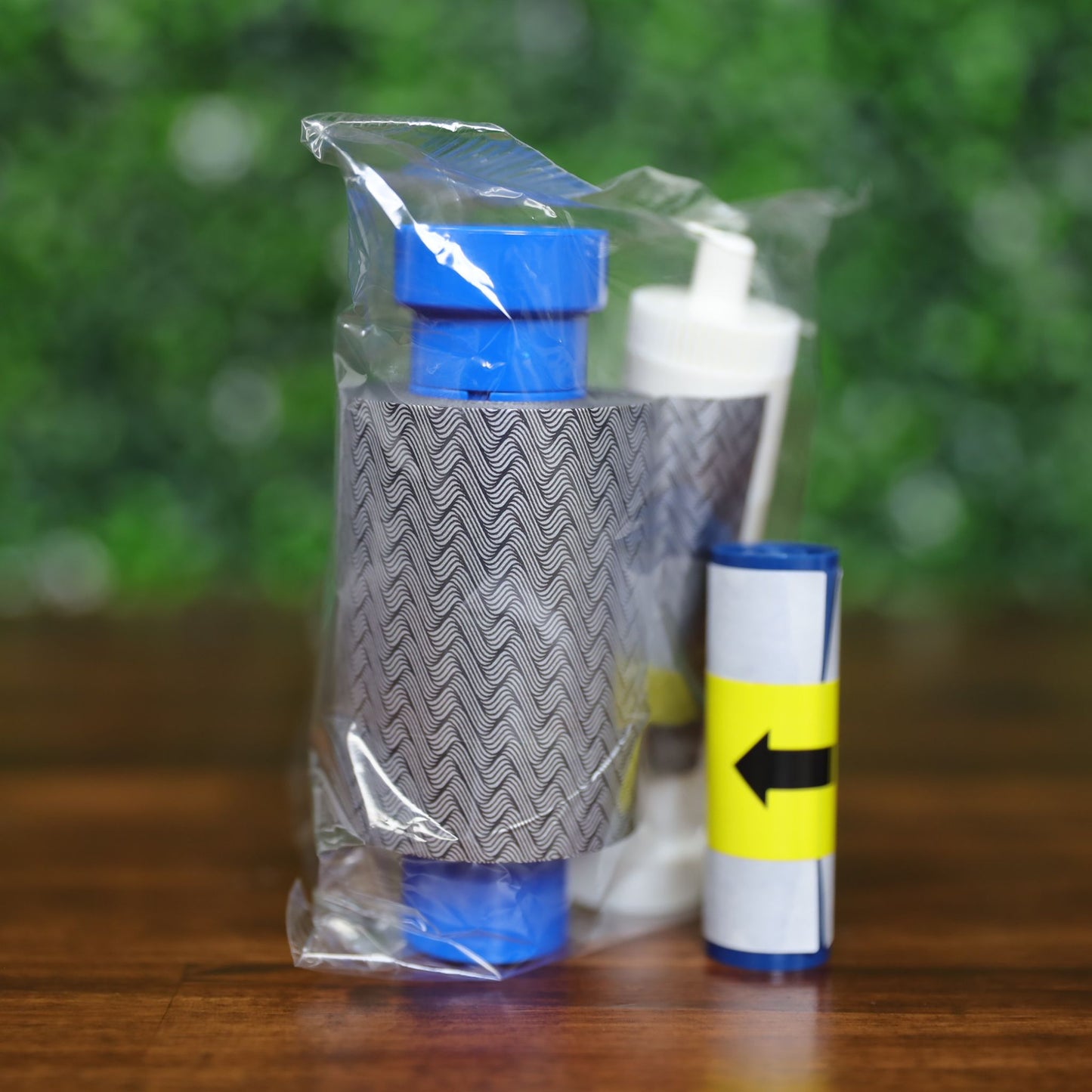 Magicard MA1000K-Scratch scratch-off ribbon with wavy gray pattern, cleaning spool, and cleaning solution bottle in clear packaging on a wood surface with green leafy background, photographed and sold by Specialist ID, an authorized Magicard dealer.