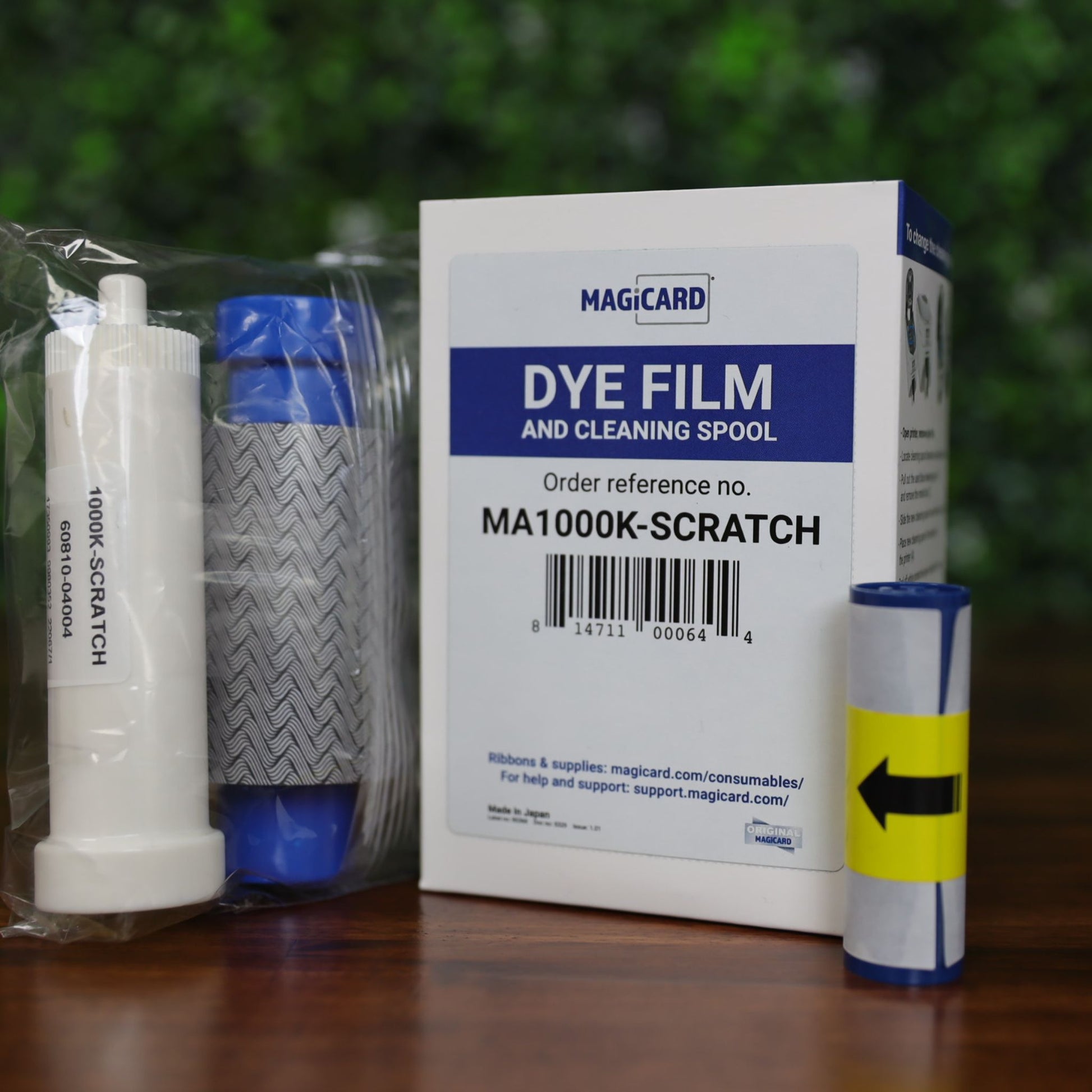 Magicard MA1000K-Scratch scratch-off ribbon kit including ribbon cartridge with wavy gray pattern, cleaning spool, and retail box, displayed on a wood surface with green leafy background, photographed and sold by Specialist ID, an authorized Magicard dealer.