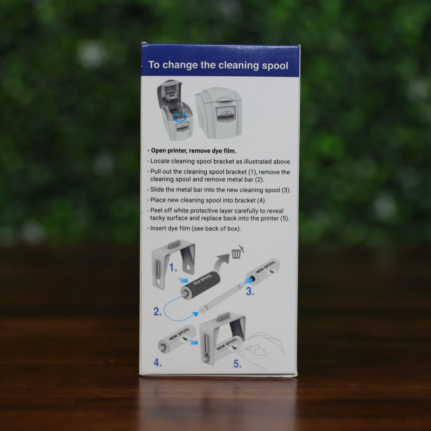 Side of Magicard MA1000K-Scratch scratch-off ribbon box showing step-by-step illustrated instructions for replacing the cleaning spool, with text and diagrams, displayed on a wood surface with green leafy background, photographed and sold by Specialist ID, an authorized Magicard dealer.