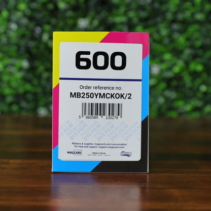 A Magicard MB250YMCKOK Color Ribbon box (250 prints) compatible with the Magicard 600 ID card printer, displayed front-facing on a wooden surface against a green blurred background, featuring the “600” label and order reference MB250YMCKOK/2 with cyan, magenta, yellow, and black accents. Photographed and sold by Specialist ID, an authorized Magicard dealer.