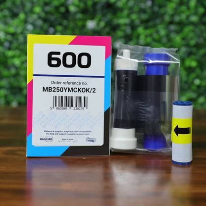 A Magicard MB250YMCKOK Color Ribbon (250 prints) compatible with the Magicard 600 ID Card Printer, shown with its box and roll, sits on a wooden surface against green foliage—perfect for your printing needs. Photographed and sold by Specialist ID, an authorized Magicard dealer.