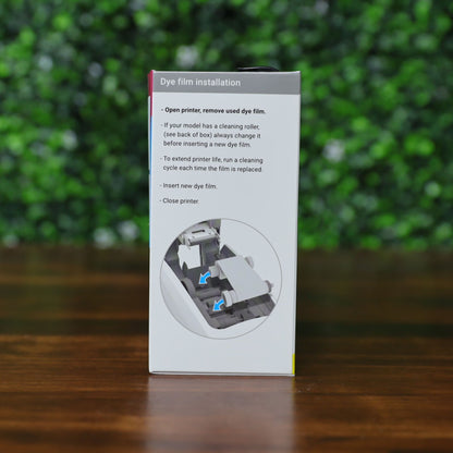 A box displaying installation instructions for the Magicard MB250YMCKOK Color Ribbon – 250 Prints, compatible with the Magicard 600 ID Card Printer, with step images and blue arrows on a blurred green background and wooden surface. Photographed and sold by Specialist ID, an authorized Magicard dealer.