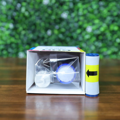 A small box containing a Magicard MB250YMCKOK Color Ribbon (250 prints, for Magicard 600 ID Card Printer) sits with a blue item and a scroll with a yellow arrow label on a wooden table against green foliage. Photographed and sold by Specialist ID, an authorized Magicard dealer.