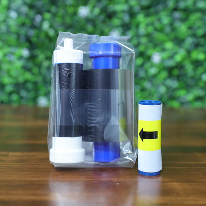 Magicard MB250YMCKOK Color Ribbons (250 prints each), for the Magicard 600 ID card printer, and a small roll with a yellow arrow label are displayed on a wooden surface against a leafy green background. Photographed and sold by Specialist ID, an authorized Magicard dealer.