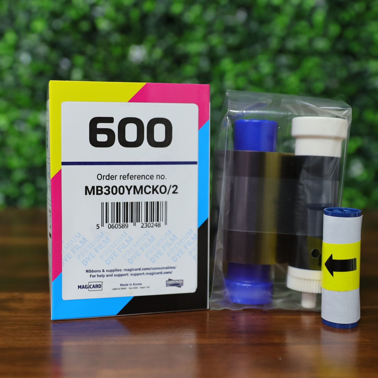 Magicard MB300YMCKO YMCKO Ribbon - 300 Prints, featuring two ribbon cartridges, is shown on a wooden surface with a green foliage background. Photographed and sold by Specialist ID, an authorized Magicard dealer.
