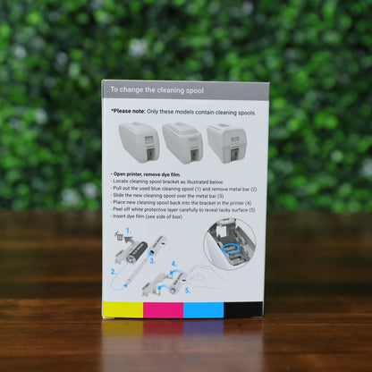 Back of a Magicard MB300YMCKOK YMCKO Ribbon box showing clear grayscale diagrams and numbered instructions with arrows for removing a used blue cleaning spool, sliding on a new spool, and placing it back into the printer bracket, with a color strip along the bottom edge, set on a wooden surface with a green leafy background. Photographed and sold by Specialist ID, an authorized Magicard dealer.