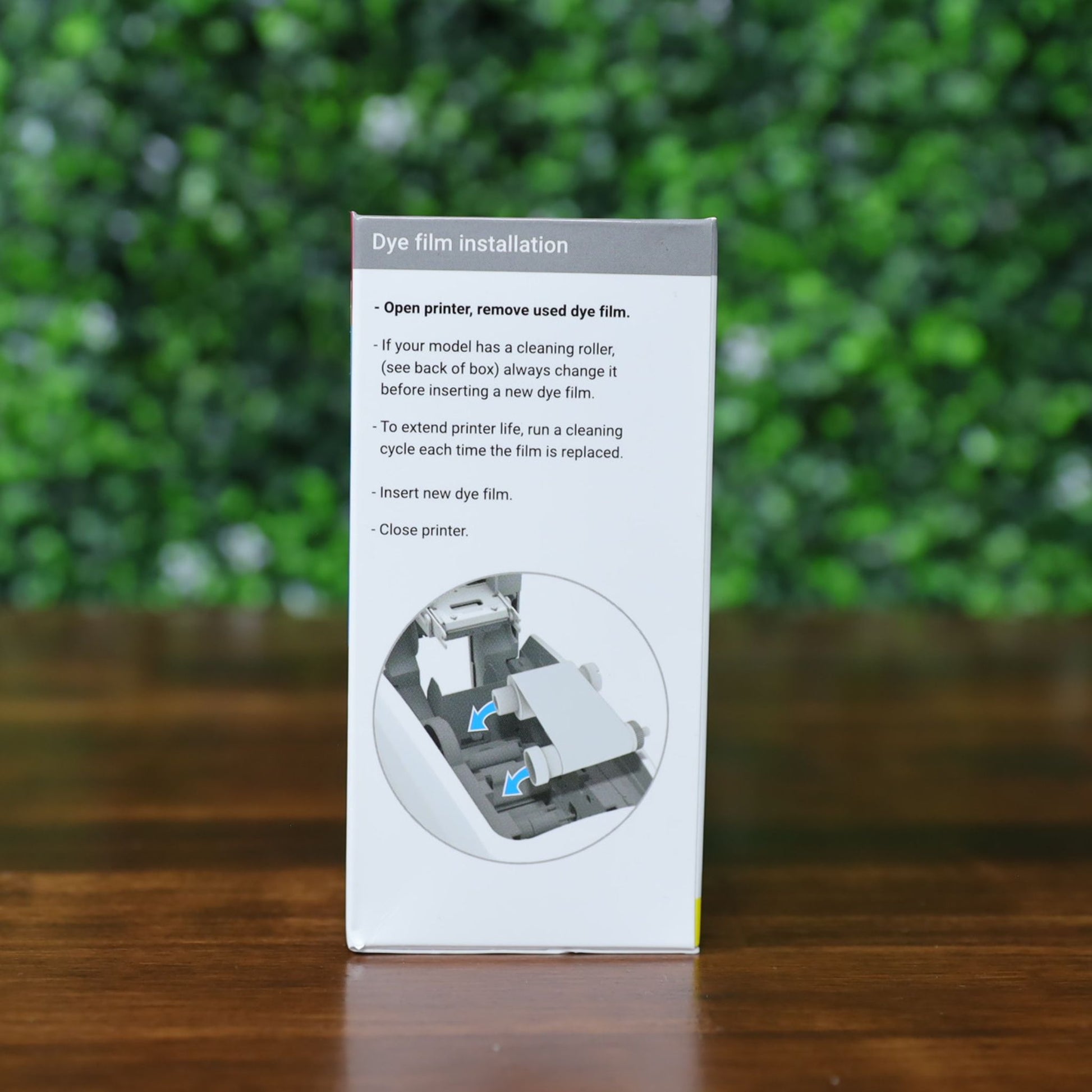 Side of a Magicard MB300YMCKOK YMCKO Ribbon box showing grayscale step-by-step instructions with arrows and a diagram on how to remove used film and insert new dye film in the printer, placed on a wooden surface with a blurred green background. Photographed and sold by Specialist ID, an authorized Magicard dealer.