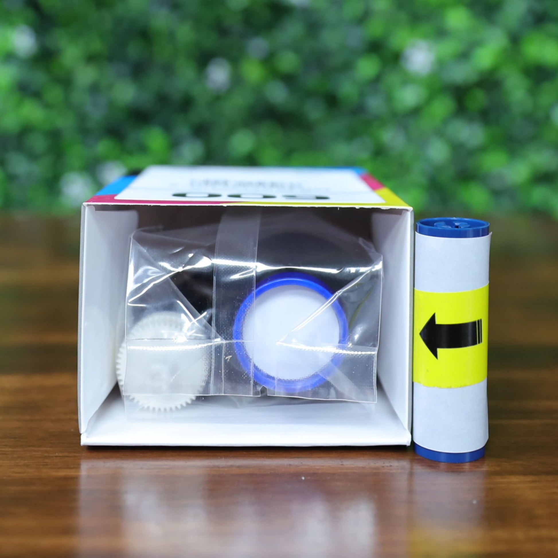 A Magicard MB300YMCKOK YMCKO Ribbon (300 prints) box lying down on its side, open to show the blue and white ribbon inside sealed in clear plastic, with a separate blue arrow cleaning roller beside it, on a wooden surface with a blurred green leafy background. Photographed and sold by Specialist ID, an authorized Magicard dealer.