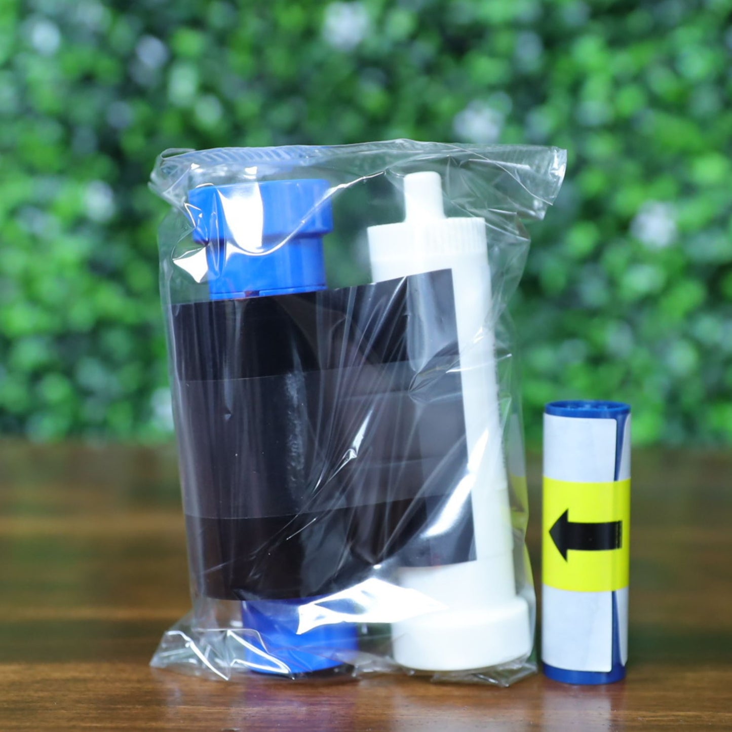 Close-up of a sealed Magicard MC600KO/2 ribbon kit showing the black overlay ribbon housed on white and blue spools, with an additional blue cleaning roller beside it. The ribbon is encased in a transparent plastic bag, resting upright on a wood surface with a blurred green background. This KO ribbon is designed for producing black resin prints with overlay protection on Magicard 600 and 300 Series card printers.
Photographed and sold by Specialist ID, an authorized Magicard dealer.