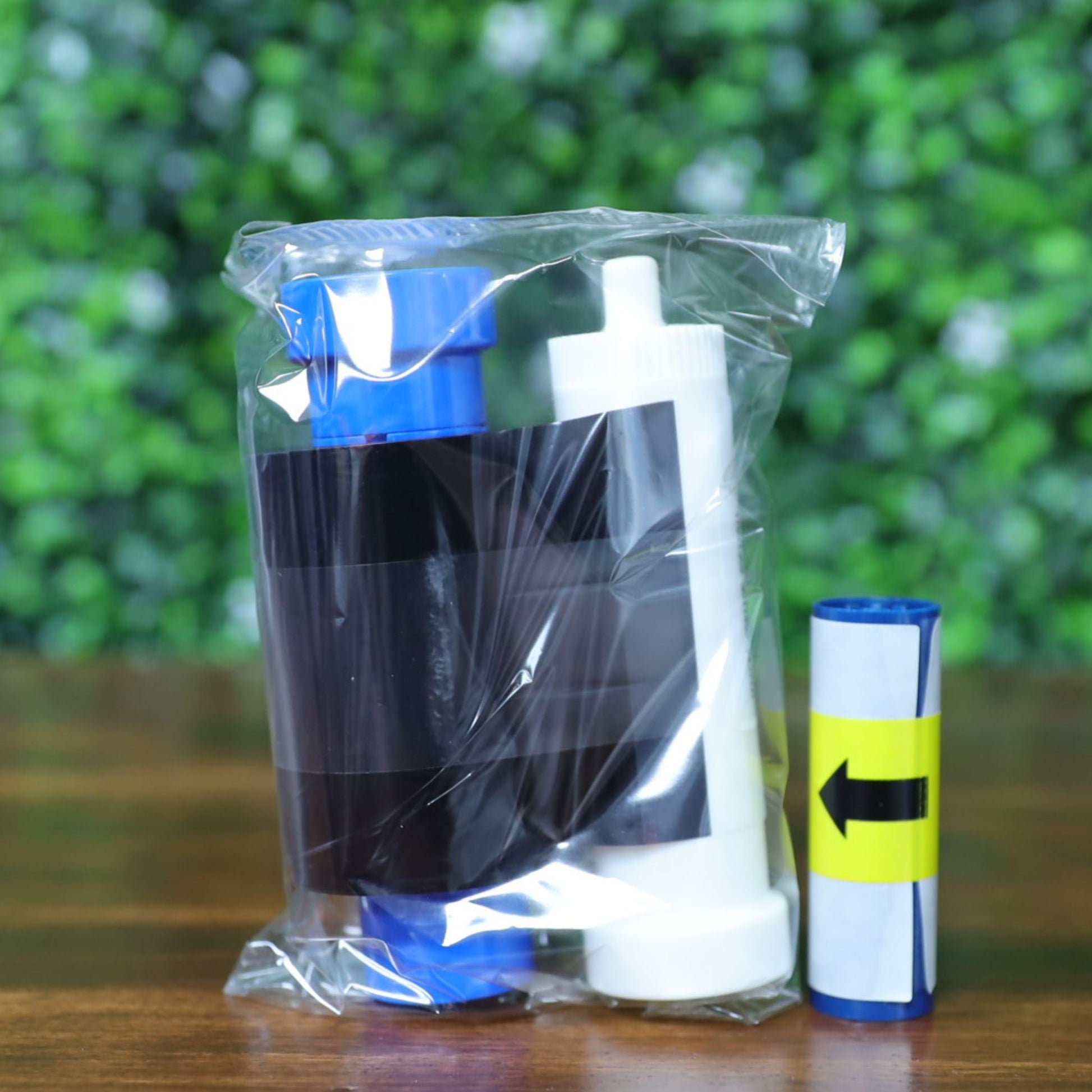 Close-up of a sealed Magicard MC600KO/2 ribbon kit showing the black overlay ribbon housed on white and blue spools, with an additional blue cleaning roller beside it. The ribbon is encased in a transparent plastic bag, resting upright on a wood surface with a blurred green background. This KO ribbon is designed for producing black resin prints with overlay protection on Magicard 600 and 300 Series card printers.
Photographed and sold by Specialist ID, an authorized Magicard dealer.