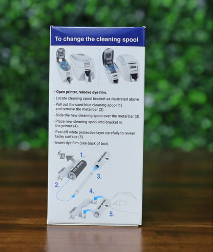 Close-up of the Magicard PR1000W ribbon box showing cleaning spool replacement instructions with illustrated steps. Includes visual diagrams of printer models, arrows indicating removal and insertion of the metal bar and cleaning spool, and labeled steps for safe installation. Photographed and sold by Specialist ID, an authorized Magicard dealer.