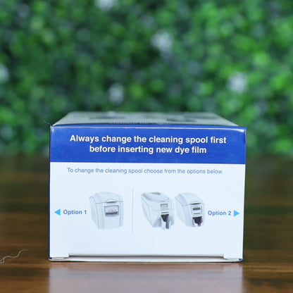 Top panel of the Magicard PR1000W dye film box showing illustrated reminder to change the cleaning spool before inserting new dye film. Includes visual options for different printer models. Photographed and sold by Specialist ID, an authorized Magicard dealer.
