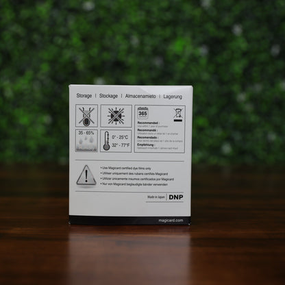 Storage instructions and certification notice on the back of a Magicard Prima433 YMCKK dye film box for 750 dual-sided ID card prints, displayed on a wood surface with a blurred green foliage backdrop. Photographed and sold by Specialist ID, an authorized Magicard dealer.