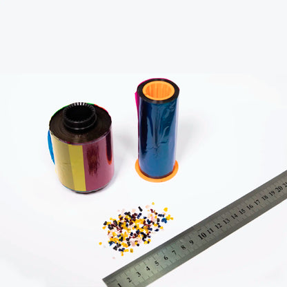 A roll of multicolored plastic film, a roll of blue plastic film on an orange spool, a pile of small colored plastic pieces, and a metal ruler placed on a white surface. The setup looks ideal for use with the Smart-Bit 651577 Ribbon Shredder by IDP to ensure data security when dealing with printer ribbons.