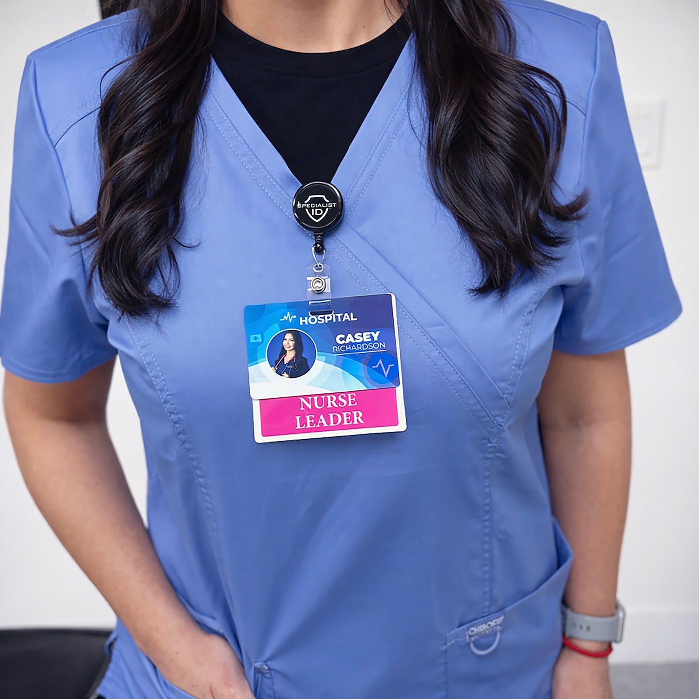 Nurse wearing a black Specialist ID badge reel clipped to blue scrubs, holding a hospital photo ID with a pink Nurse Leader badge buddy