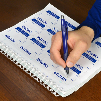 A person is meticulously writing on a sheet of Expiring Visitor Badge and Log Book - 480 Badges (05741) using a blue pen, ensuring accurate entries for the visitor management system.