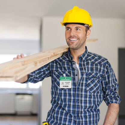 A construction worker in a yellow hard hat and plaid shirt carries a wooden beam on his shoulder and smiles. He wears a visible Preprinted Self-Expiring Badge Backpart, Box of 1000 (P/N T59XX).