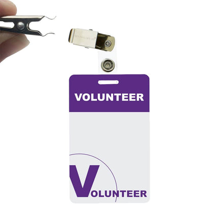 Hand placing a clip-on badge holder onto a white and purple name tag labeled "Volunteer" with 5 Pack - Purple Heavy Duty Plastic Volunteer Badges with Clothing Friendly Clip, ensuring it is clothing friendly.