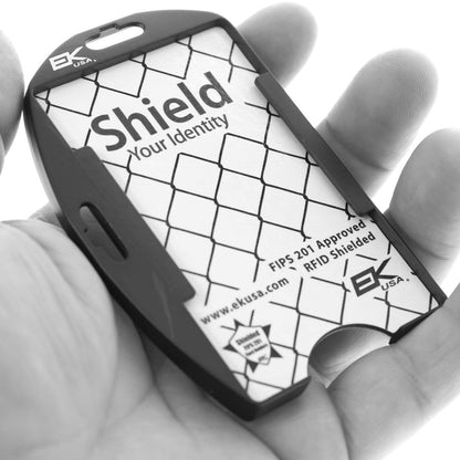 A hand holds a Black EK Patriot Shielded RFID Blocking Two Card ID Badge Holder (10916) by EK USA designed to protect identity cards from electronic theft, with the text "Shield Your Identity" and "FIPS 201 Approved." The product has a lattice pattern and is perfect for government-issued CAC cards.