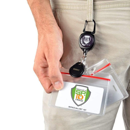 A person holds a Heavy Duty Clear Vinyl Horizontal Badge Holder With Resealable Zip Top (1815-1010) attached to a retractable keychain clipped to their pants. The badge displays the logo and text "SPECIALIST ID INC.