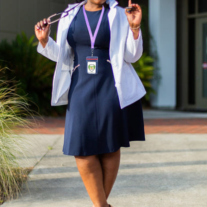 Person in a navy dress and white coat, holding glasses and a stethoscope. They are wearing a lanyard with a Clear Vertical Vinyl ID Holder with a Zipper Top (VBH-V-ZIP) and standing outside on a sunny day.