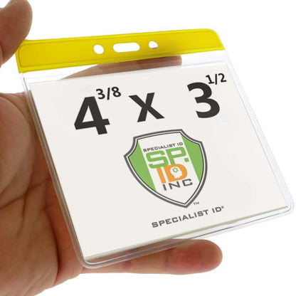 A hand holds an Extra Large Color Code Badge Holder for 4x3 Badges - Horizontal Vinyl with Color Bar (1820-120X) with a yellow top, displaying a card that reads "4 3/8 x 3 1/2" and features the "Specialist ID Inc" logo.