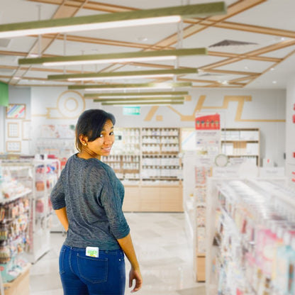 A woman in a gray top and blue jeans stands in a brightly lit store aisle, looking back over her shoulder. The store shelves are stocked with various items, including Clear Vinyl Business Card Holder - Medicare Card Protective Sleeve (1840-3505) neatly organized by the register.
