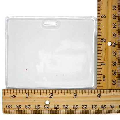 Clear plastic ID badge holder placed next to a wooden ruler for scale; this Clear Flexible Vinyl Horizontal Proximity Card Holder (P/N 1840-5010), with dimensions approximately 3.5 inches by 2.5 inches, features a top load design for easy use.