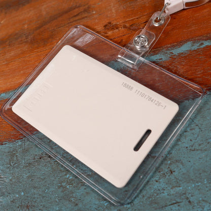 A white ID card in a Clear Heavy Duty Vinyl Horizontal Proximity Card Holder 1840-5017 with a metal clip is placed on a wooden surface. The horizontal proximity card holder shows a numerical code and has a slot for attachment.