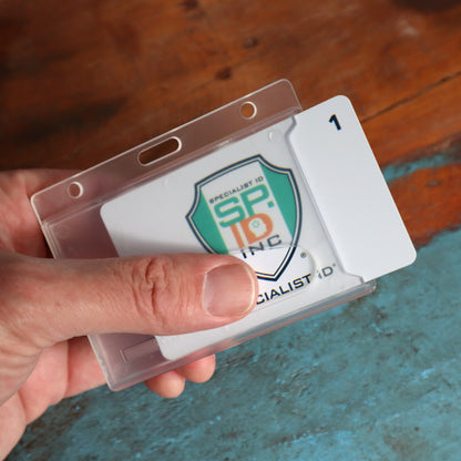 A hand holding a Horizontal Rigid Plastic Badge Holder - Frosted Card Dispenser (P/N 1840-6000) with a card inside displaying a green and orange shield logo and the text "SPECIALIST ID INC." on a wooden surface background, showcases excellent ID badge protection.