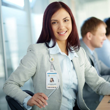 A woman in professional attire smiles while sitting at a desk, her ID badge securely clipped to a Vertical / Horizontal Durable Clear Vinyl ID Badge Holder (1815-1600). Other workers are visible in the professional workplace environment, creating a bustling yet focused atmosphere.