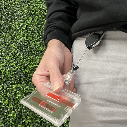 A person holds a retractable badge holder attached to their waistband, featuring a 2 Card Badge Holder with Slide (P/N 1840-6400) with a red extractor slide. In hand, they have a clear plastic ID card case. The background showcases lush green foliage.