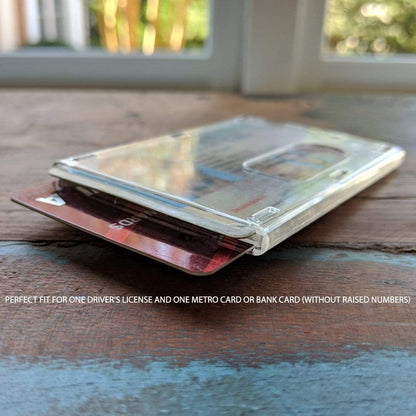 A close-up of a transparent Rigidwear Clear Vertical 2-Card Badge Holder 1840-6560 (Jam P/N 706-NN2) placed on a wooden surface, containing a driver's license and a Metro Card, with text stating it fits a driver's license and a card without raised numbers.