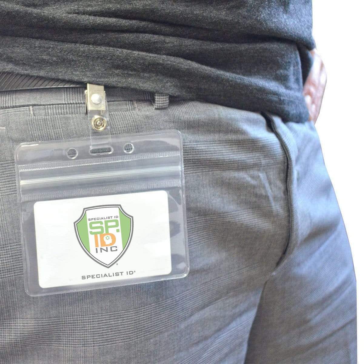 A close-up image of a person wearing gray pants and a black shirt, with a clear plastic badge holder featuring an ID Badge Strap Clips (Industry Standard Clip) attached to their belt. The badge holder contains a card displaying the logo "Specialist ID Inc.