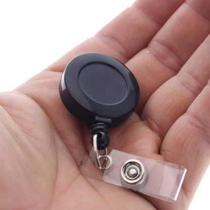 A close-up of a hand holding a Badge Reel with Belt Clip (P/N 2120-303X) with a clear plastic strap, metal snap, and an integrated belt clip.
